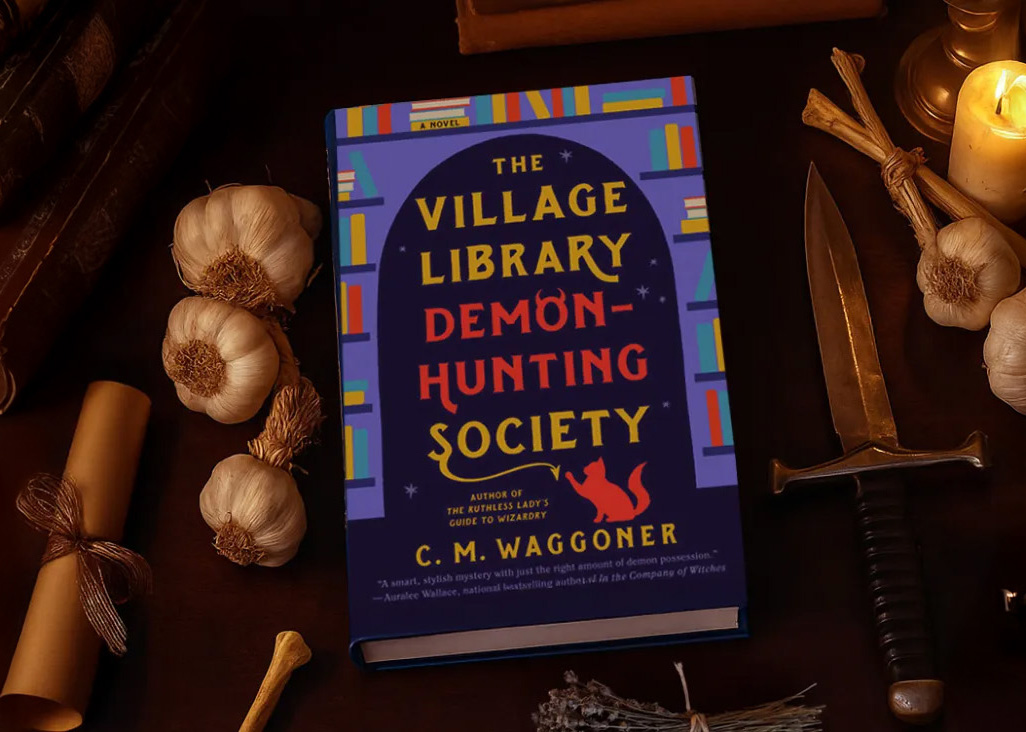 Overhead view of the book "The Village Library Demon-Hunting Society" atop a table next to a rope of garlic and dagger.