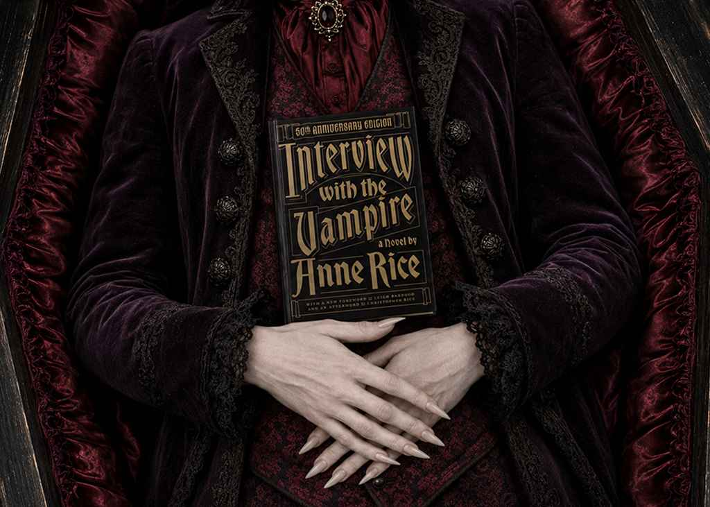 A pale vampire with long nails lies in a coffin, holding the 50th anniversary edition of Interview with the Vampire against a dark red and purple gothic outfit.